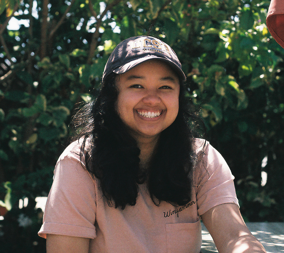 A headshot of myself sitting in front of plants wearing a navy cap (Go Gauchos!) and pink shirt